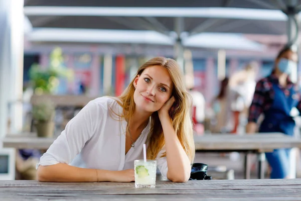 Молодая женщина пьет коктейль мохито на террасе кафе в жаркий летний день. Красивая деловая женщина наслаждается теплым вечером в ресторане после работы. Счастливая улыбающаяся одинокая леди. — стоковое фото