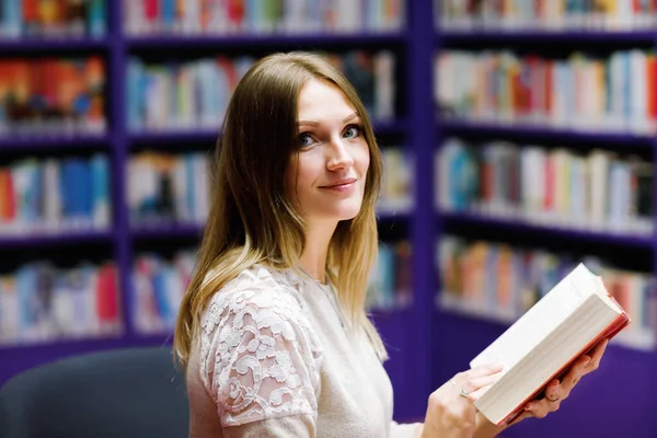 Young beautiful woman in public library, holding, reading and choosing books for leisure or education. Woman making research for college study — Stock Photo, Image