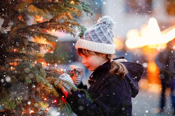Niña linda que se divierte en el mercado tradicional de Navidad durante las fuertes nevadas. Niño feliz disfrutando del mercado familiar tradicional en Alemania. Colegiala de pie junto al árbol de Navidad iluminado. — Foto de Stock