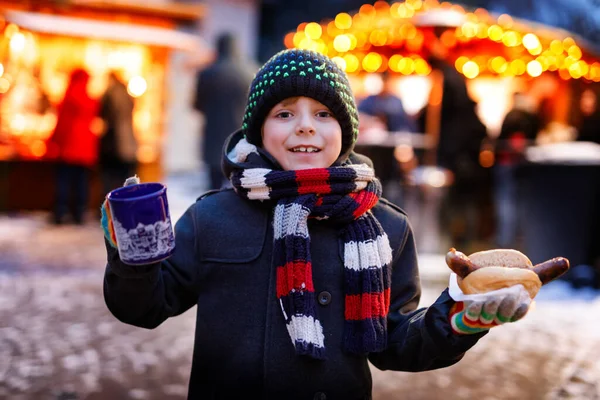 Little cute kid boy eating German sausage and drinking hot children punch on Christmas market. Happy child on traditional family market in Germany. Laughing boy in colorful winter clothes — Stock Photo, Image