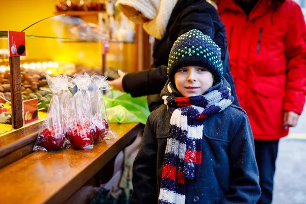 Kleine süße Junge in der Nähe von süßen Stand mit gezuckerten Äpfeln und Schokoladenfrüchten. Glückskind auf dem Weihnachtsmarkt in Deutschland. Traditionelle Freizeit für Familien an Weihnachten. Feiertag, Feier, Tradition. — Stockfoto