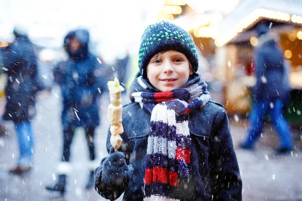在传统的德国圣诞市场上，可爱的小男孩吃着白色的巧克力，上面涂满了扭曲的水果。雪天期间，德国传统家庭市场上的快乐孩子. — 图库照片