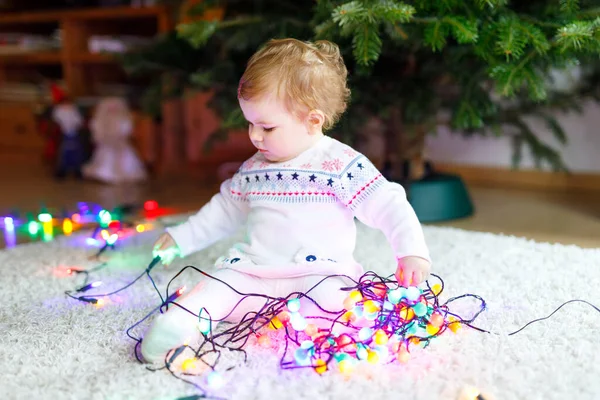 可愛い手でカラフルなライトガーランドを保持する愛らしい赤ちゃんの女の子。クリスマスツリーを家族で飾るお祝い服の小さな子供。Weihnachtenと呼ばれる伝統的な休日の最初のお祝い — ストック写真