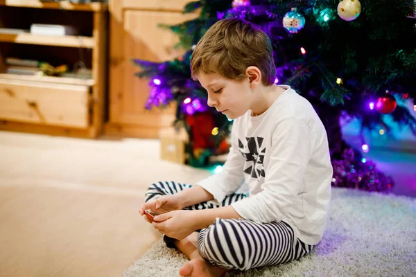 Cute little blond kid boy using smartphone on Christmas with decorated tree on background. Happy healthy hild having fun at home. Popular gift for xmas — Stock Photo, Image