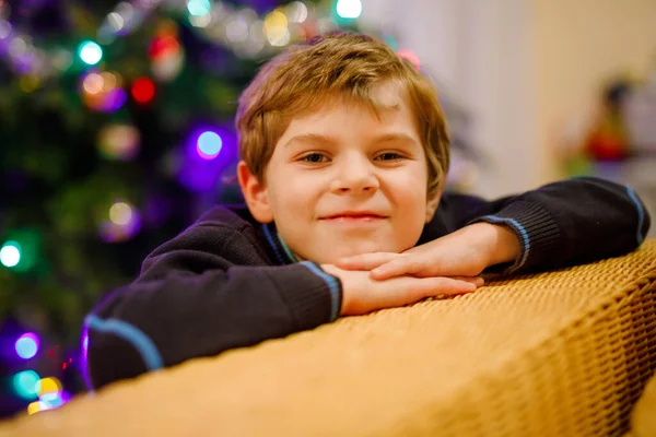 Liten pojke på julafton med julgran och ljus på bakgrunden. Med färgglad belysning och krans. Glad förskolebarn firar jul, familjesemester. Pojke i nattkläder. — Stockfoto