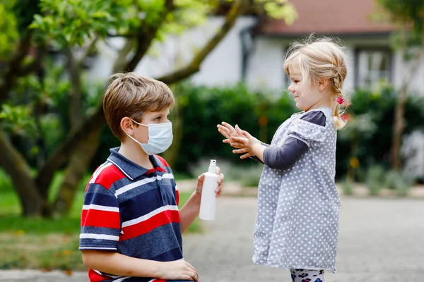 Niño de la escuela ayudando a la hermana pequeña a limpiarse las manos con aerosol desinfectante. Hermano y linda niña aprenden reglas de higiene. Familia durante el cierre del coronavirus y la gripe. Protección del virus covid. — Foto de Stock