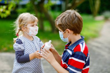 Okul çocuğu küçük kız kardeşine tıbbi maske takması için yardım ediyor. Sanitzier spreyi tutan kız. Kardeş ve tatlı küçük kız hijyen kurallarını öğreniyorlar. Coronavirus kapanışı sırasında aile. Kapsamlı koruma.