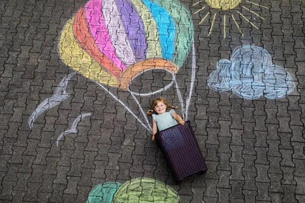 Glückliches kleines Mädchen, das im Heißluftballon fliegt und im Sommer mit bunten Kreiden in Regenbogenfarben auf Boden oder Asphalt gemalt wird. Nettes Kind, das Spaß hat. Kreative Freizeit für Kinder. — Stockfoto