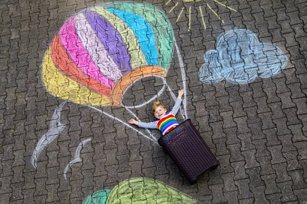 Felice bambina che vola in mongolfiera dipinta con gessetti colorati in colori arcobaleno a terra o asfalto in estate. Bel bambino che si diverte. svago creativo per bambini. — Foto Stock