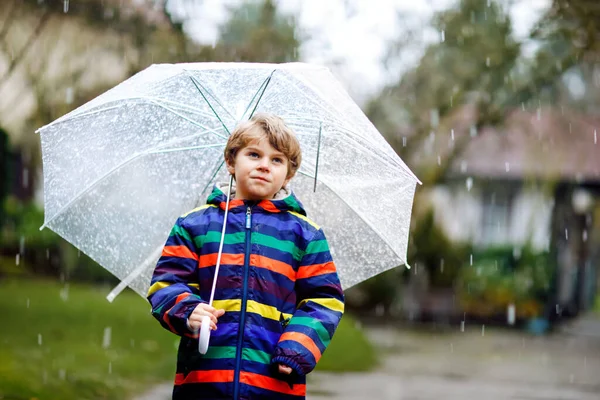 寒い日に傘を持って学校に行く途中の美しい小さな男の子は、雨や雪の中を歩いています。カラフルなファッションで幸せと喜びの子供カジュアル服. — ストック写真