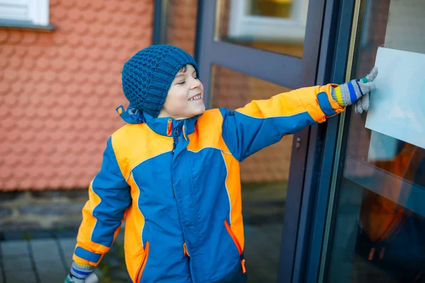 En liten skolpojke i grundskolan som går till skolan en kall vinterdag. Glada barn i varma vinterkläder knuffar dörren till skolan, utomhus. — Stockfoto