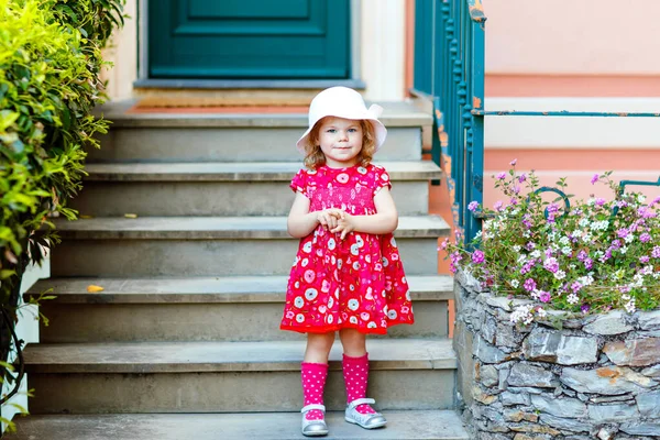 Porträtt av vacker liten gorgeus härlig småbarn flicka i rosa sommar titta kläder, mode klänning, knästrumpor och hatt. Glad frisk baby barn poserar framför färgglada hus. — Stockfoto