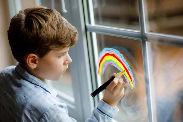 Bella scuola ragazzo in pigiama pittura arcobaleno con il colore della finestra colorato durante la quarantena pandemica coronavirus. Bambini che dipingono arcobaleni in tutto il mondo con le parole Lascia che tutto vada bene. — Foto Stock