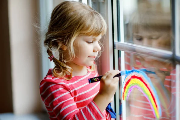 Adoralbe liten småbarn flicka med regnbåge målad med färgglada fönster färg under pandemisk coronavirus karantän. Barnmålning regnbågar och hjärtan runt om i världen med ord Låt alla må bra. — Stockfoto