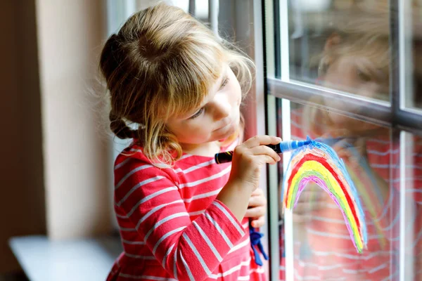 Adoralbe petite fille tout-petit avec arc-en-ciel peint avec couleur de fenêtre colorée pendant la quarantaine de coronavirus pandémique. Enfant peignant des arcs-en-ciel et des cœurs autour du monde avec des mots Soyons tous bien. — Photo