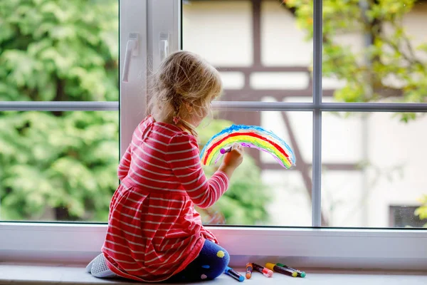 Adoralbe kleine peuter meisje met regenboog geschilderd met kleurrijke venster kleur tijdens pandemische coronavirus quarantaine. Kind schilderen regenbogen en harten over de hele wereld met woorden Laten we allemaal goed zijn. — Stockfoto