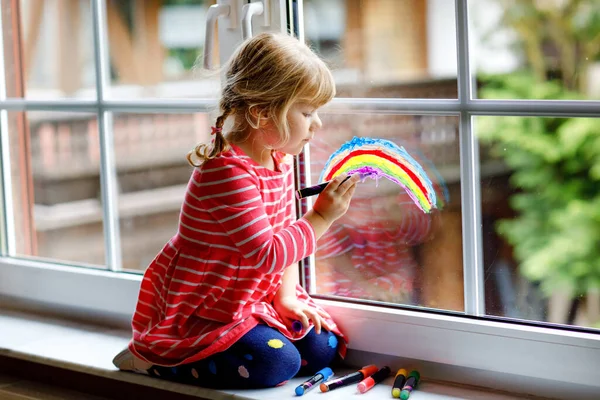 Adoralbe petite fille tout-petit avec arc-en-ciel peint avec couleur de fenêtre colorée pendant la quarantaine de coronavirus pandémique. Enfant peignant des arcs-en-ciel et des cœurs autour du monde avec des mots Soyons tous bien. — Photo