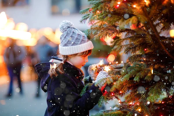 Malá roztomilá holčička baví na tradičním vánočním trhu během silného sněžení. Šťastné dítě těší tradiční rodinný trh v Německu. Školačka stojící ozářený vánoční strom. — Stock fotografie