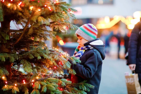 Pequeño niño lindo que se divierte en el mercado tradicional de Navidad durante las fuertes nevadas. Niño feliz disfrutando del mercado familiar tradicional en Alemania. Colegial de pie junto al árbol de Navidad iluminado. —  Fotos de Stock