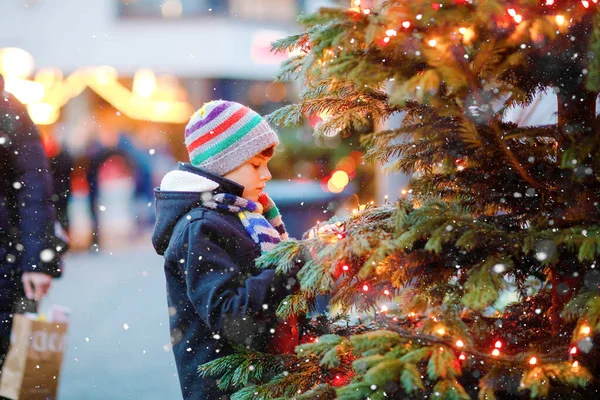 Pequeño niño lindo que se divierte en el mercado tradicional de Navidad durante las fuertes nevadas. Niño feliz disfrutando del mercado familiar tradicional en Alemania. Colegial de pie junto al árbol de Navidad iluminado. — Foto de Stock