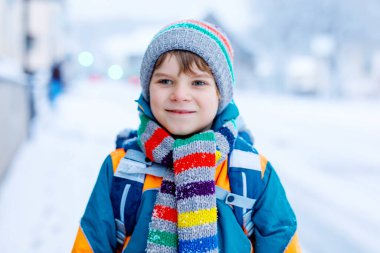 Şiddetli kar yağışı sırasında okula yürüyen ilkokul çocuğu. Sabahın erken saatleri ve şehirde karlı sokaklar. Sırt çantalı ya da renkli kış kıyafetleri içinde bir çocuk.