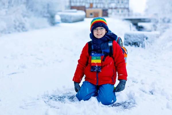 Underbar liten skolpojke med glasögon av elementär klass som går till skolan under starkt snöfall. Snöiga gator i stan. Barn med ryggsäck eller väska i färgglada vinterkläder — Stockfoto