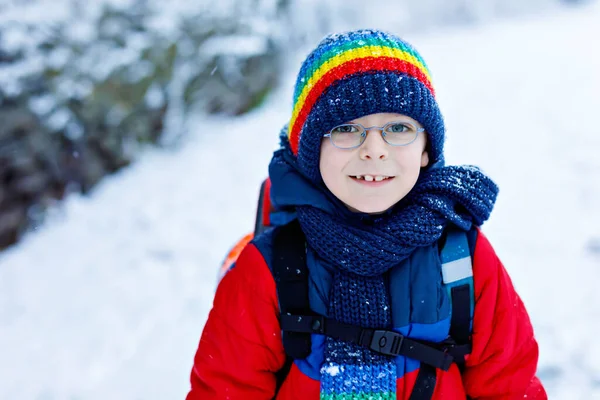 豪雪の中、小学生の眼鏡をかけて学校まで歩くゴージャス小学生少年。街の雪の通り。カラフルな冬服のバックパックやサッチェルを持つ子供 — ストック写真
