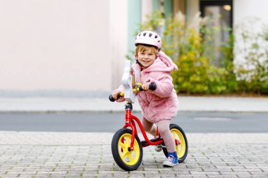 Kask takıp kreşe, çocuk parkına ya da çocuk parkına giden dengeli bisiklete binen tatlı küçük kız. Mutlu çocuk bisiklet sürerek öğrenirken eğleniyor. Soğuk bir sonbahar gününde açık havada hareketli bir çocuk..
