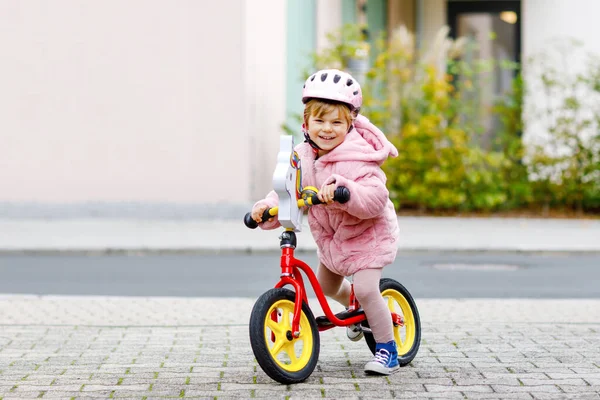 Cute little toddler girl with helmet riding on run balance bike to daycare, playschool or kindergarden. Happy child having fun with learning on learner bicycle. Active kid on cold autumn day outdoors. — Stock Photo, Image