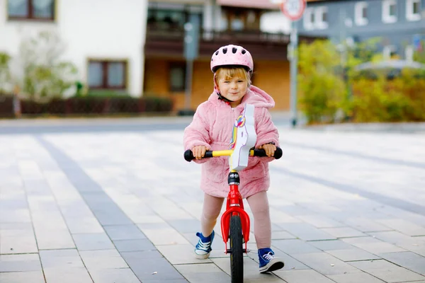 Linda niña con casco montado en bicicleta de equilibrio de carrera a la guardería, escuela de juegos o jardín de infantes. Niño feliz divirtiéndose con el aprendizaje en bicicleta de aprendizaje. Niño activo en el frío día de otoño al aire libre. — Foto de Stock