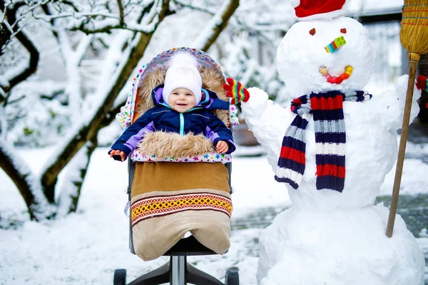 Linda niñita hermosa sentada en el cochecito o cochecito en el día de invierno con nieve y muñeco de nieve — Foto de Stock