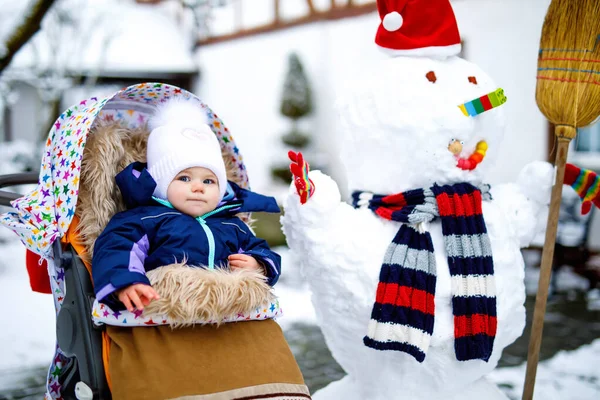 Bonito menina bonita sentada no carrinho de bebê ou carrinho no dia de inverno com neve e boneco de neve — Fotografia de Stock