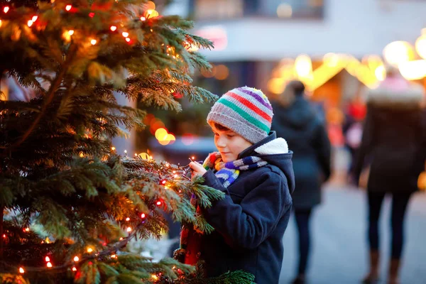 Little cute kid boy having fun on traditional Christmas market during strong snowfall. Happy child enjoying traditional family market in Germany. Schoolboy standing by illuminated xmas tree. — Stock Photo, Image