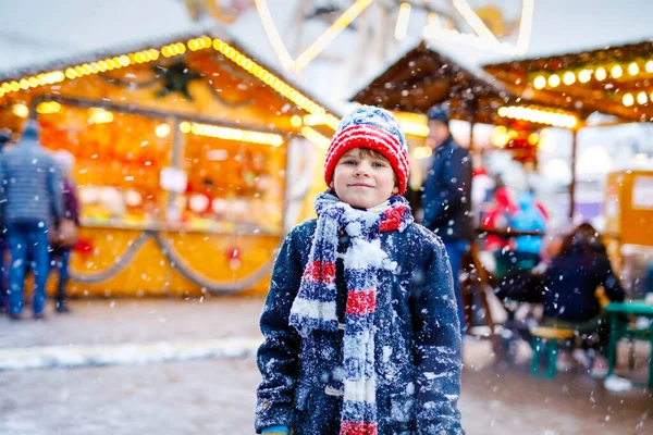 Little cute kid boy having fun on traditional German Christmas market during strong snowfall.. Happy child enjoying traditional family market in Germany, Munich. Laughing boy in colorful clothes — Stock Photo, Image