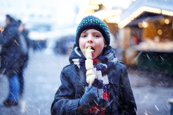 Kleine schattige jongen eten witte chocolade bedekt fruit op spiesje op traditionele Duitse kerstmarkt. Gelukkig kind op traditionele familiemarkt in Duitsland tijdens sneeuwdag. — Stockfoto