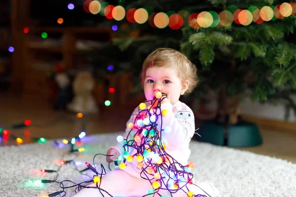 Schattige baby meisje met kleurrijke lichten bloemenslinger in schattige handen. Klein kind in feestelijke kleding versieren kerstboom met familie. Eerste viering van de traditionele feestdag genaamd Weihnachten — Stockfoto