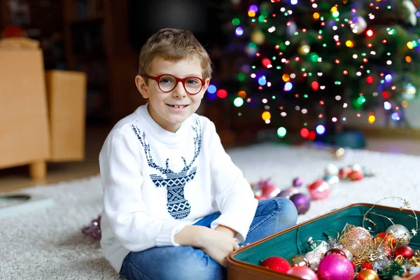 Schöner kleiner Junge mit Brille und buntem Vintage-Weihnachtsspielzeug und Bällen im alten Koffer. Kind schmückt Weihnachtsbaum — Stockfoto