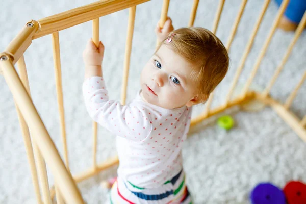 Beautiful little baby girl standing inside playpen. Cute adorable child playing with colorful toys. Home or nursery, safety for kids. Alone baby waiting for mom — Stock Photo, Image