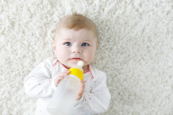 Симпатичная маленькая упрямая девочка держит бутылочку для кормления и пьет молочные смеси. Первая еда для детей. Новорожденный ребенок, маленькая девочка лежит на белом фоне. Семья, новая жизнь, детство, бутылка — стоковое фото