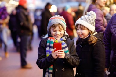 Two little kids, boy and girl having fun on traditional Christmas market during strong snowfall with taking photos with mobile smartphone or phone. Happy children, siblings and best friends clipart