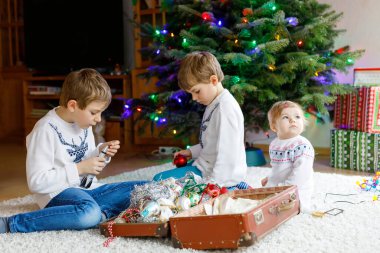 İki küçük oğlan ve sevimli kız bebek Noel ağacını eski model oyuncaklar ve toplarla süslüyorlar. Aile tatili için aile hazırlık kutlaması. Evde üç çocuk, erkek ve kız kardeş var..