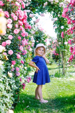 Çiçek açan gül bahçesinde küçük bir kızın portresi. Yaz güneşli bir günde parkta güllerle ve çiçeklerle eğlenen sevimli güzel bir çocuk. Mutlu gülümseyen bebek.