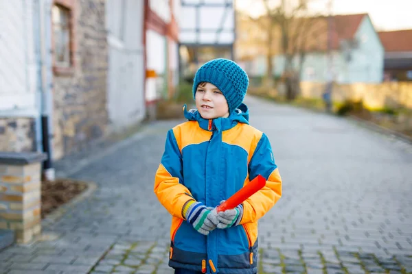 Niño de la escuela primaria caminando a la escuela en el frío día de invierno. Niño feliz divirtiéndose en una calle de la ciudad. Estudiante con mochila en ropa de invierno colorida sosteniendo cosas para el proyecto —  Fotos de Stock