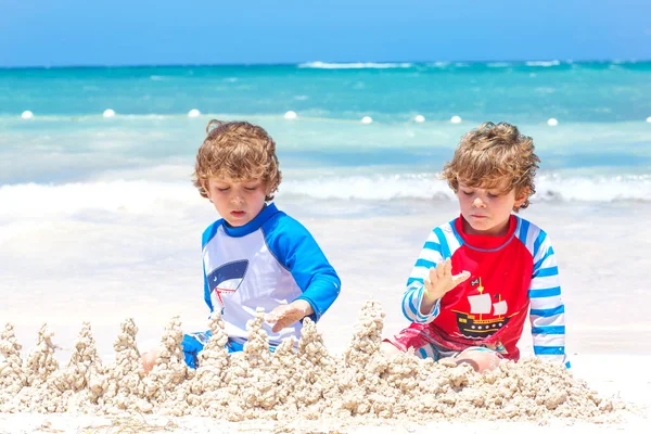 Dos niños divirtiéndose con la construcción de un castillo de arena en la playa tropical en la isla. Niños sanos jugando juntos en sus vacaciones. Gemelos, hermanos felices riendo y sonriendo —  Fotos de Stock