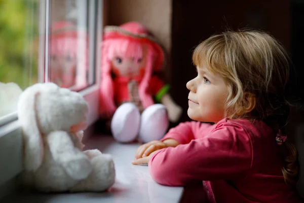 Petite fille mignonne assise à côté de la fenêtre et regardant dehors le jour de pluie. Enfant rêveur avec poupée et jouet doux se sentant heureux. Self isolation concept during corona virus pandemic time. Enfant solitaire. — Photo