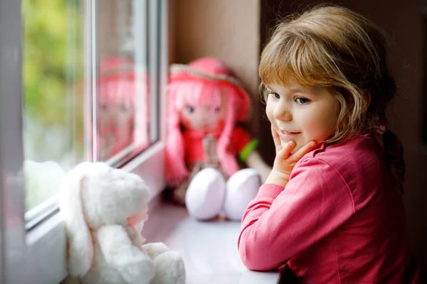 Симпатичная малышка сидит у окна и смотрит в дождливый день. Мечтающий ребенок с долой и мягкой игрушкой чувствует себя счастливым. Концепция самоизоляции во время пандемии коронного вируса. Одинокий ребенок. — стоковое фото