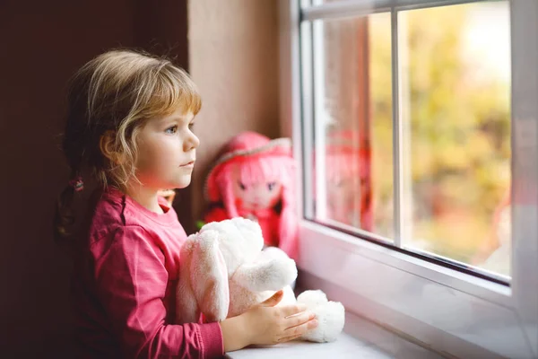 Schattig peuter meisje zit bij het raam en kijkt uit op regenachtige dag. Dromend kind met pop en knuffel die zich gelukkig voelen. Zelfisolatieconcept tijdens de corona-viruspandemie. Eenzame jongen. — Stockfoto