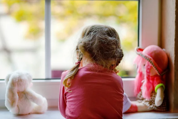 Petite fille mignonne assise à côté de la fenêtre et regardant dehors le jour de pluie. Enfant rêveur avec poupée et jouet se sentant seul. Self isolation concept during corona virus pandemic time. sans visage, méconnaissable — Photo