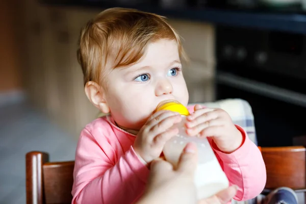 Jolie adorable petite fille tenant le biberon et le lait maternisé. Première nourriture pour bébés. Enfant nouveau-né, assis dans la chaise de la cuisine domestique. Bébés en santé et concept d'alimentation au biberon — Photo