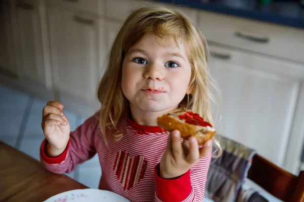 Linda niña divertida come bollo dulce para el desayuno. Feliz niño comiendo panecillo con mermelada de fresa. Alimento saludable para niños y niños con gelatina hecha a sí mismos — Foto de Stock